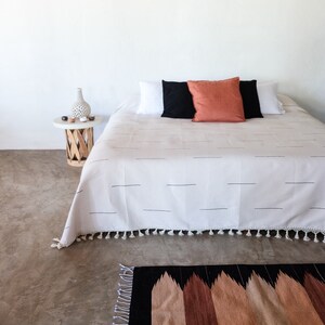 Puede incluir: Una cama blanca con una manta de rayas blancas y negras y una almohada roja. La cama est&aacute; en una habitaci&oacute;n con una pared blanca y una mesita de noche de madera con un jarr&oacute;n y algunos objetos peque&ntilde;os encima. Hay una alfombra de patrones negros y marrones delante de la cama.