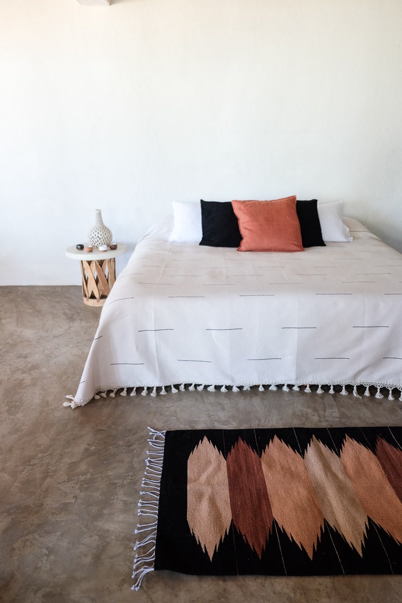 Puede incluir: Una cama blanca con una manta de rayas blancas y negras y una almohada roja. La cama est&aacute; en una habitaci&oacute;n con una pared blanca y una mesita de noche de madera con un jarr&oacute;n y algunos objetos peque&ntilde;os encima. Hay una alfombra de patrones negros y marrones delante de la cama.