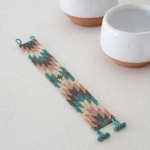 May include: A beaded bracelet with a geometric pattern in shades of teal, pink, and brown. The bracelet has a fringe on each end.