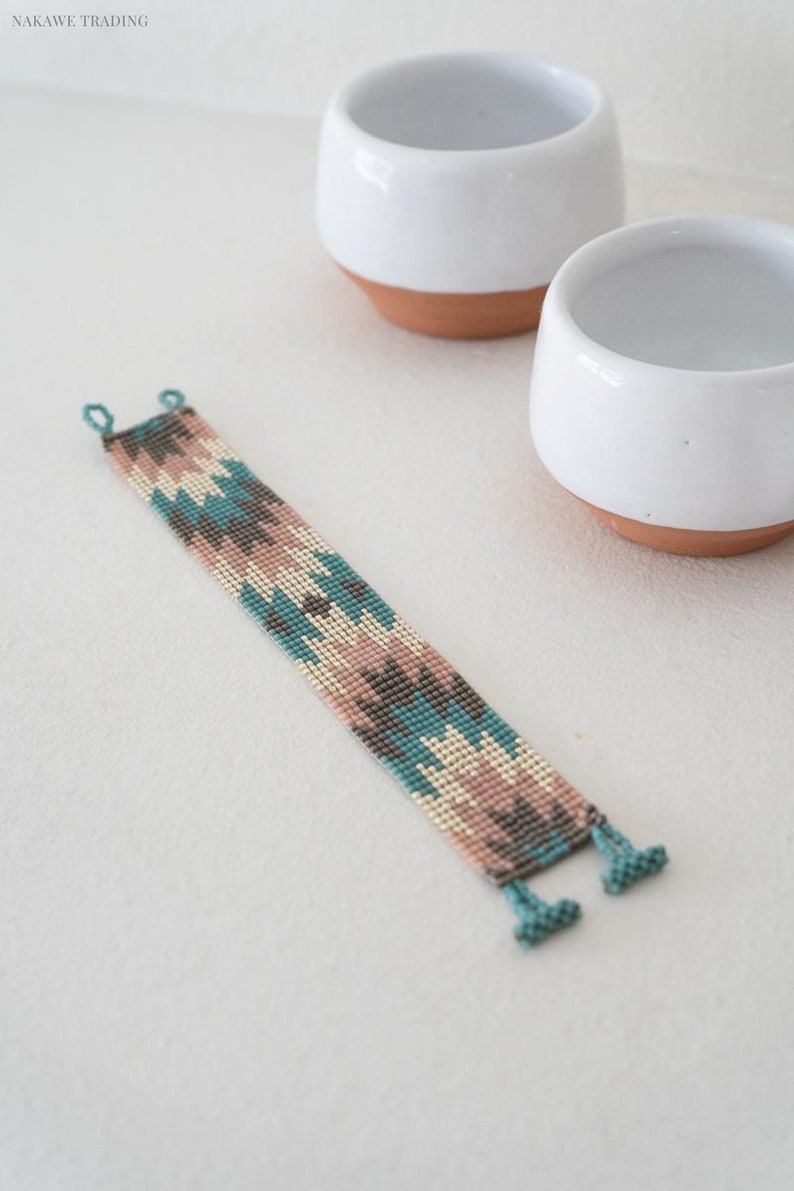 May include: A beaded bracelet with a geometric pattern in shades of teal, pink, and brown. The bracelet has a fringe on each end.