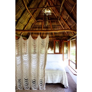 May include: A white macrame curtain hangs in front of a bed with white linens and a thatched roof. The bed is in a room with a wooden floor and a window with a view of the outdoors.