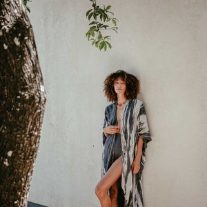 May include: A woman wearing a long, flowing, grey and white patterned kimono with a deep V-neckline. She is standing in front of a white wall with a tree branch hanging down.