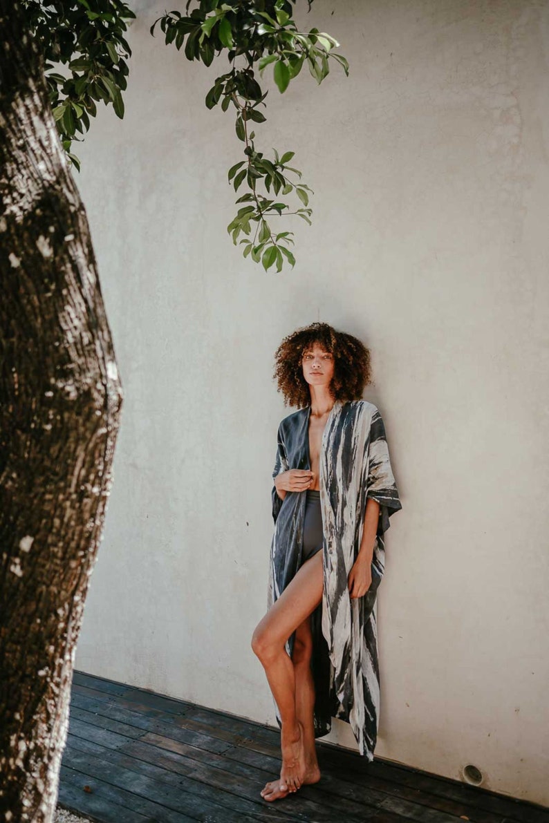 May include: A woman wearing a long, flowing, grey and white patterned kimono with a deep V-neckline. She is standing in front of a white wall with a tree branch hanging down.