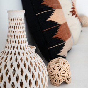 May include: Two white ceramic vases with intricate cut-out designs. One vase is tall and slender, the other is round and squat. Both vases have a light brown color.