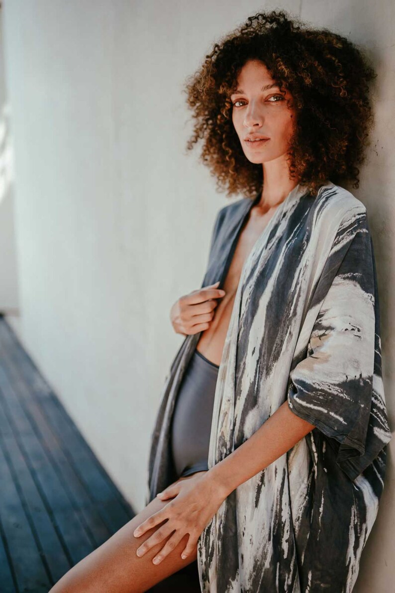 May include: A woman wearing a grey and white patterned kimono with a black swimsuit underneath. The kimono has a loose fit and is open at the front, revealing the swimsuit.