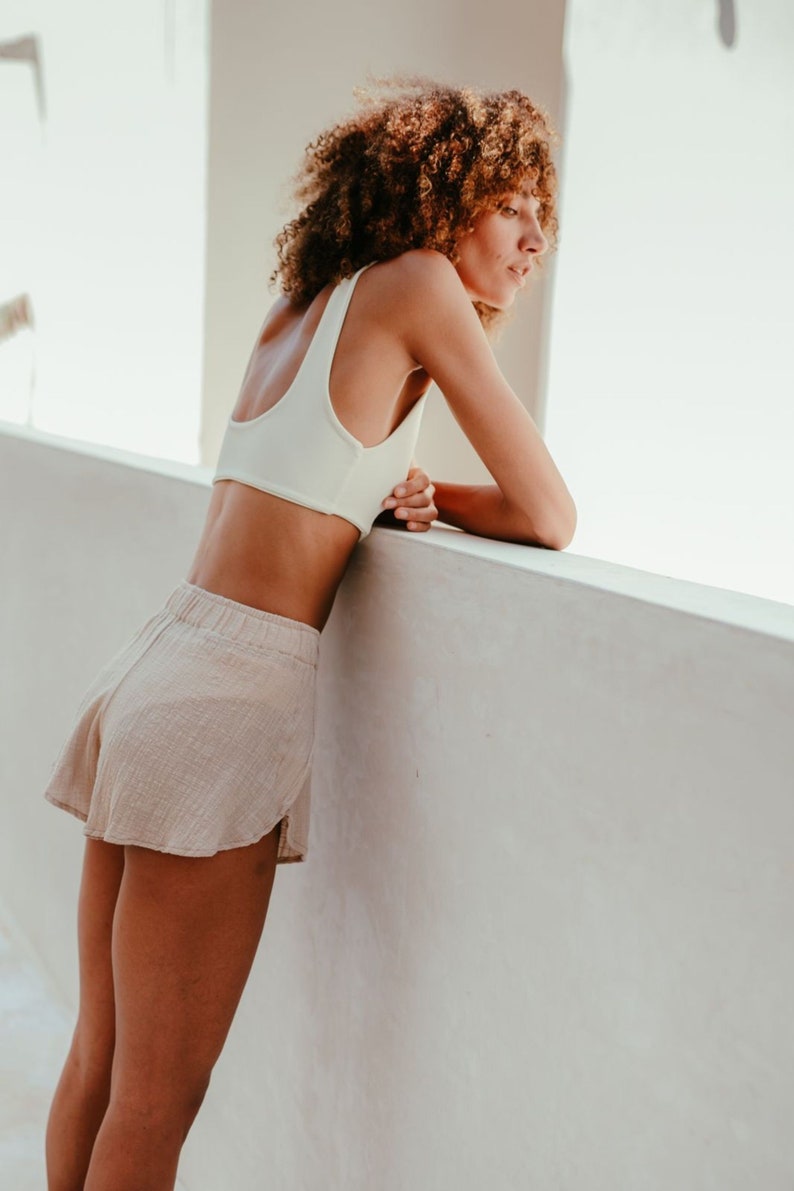 May include: A woman wearing a white crop top and beige linen shorts leans against a white wall.