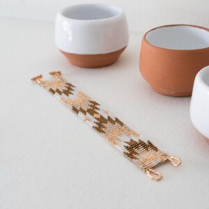 May include: A white and brown beaded bracelet with a geometric pattern. The bracelet is made with small, evenly spaced beads and has gold-toned clasps.