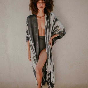 May include: A woman wearing a long, grey and white patterned kimono with a black high-waisted swimsuit bottom. She is standing in front of a light beige wall.