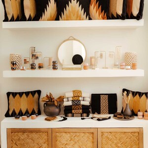 May include: A white shelf unit with two shelves, featuring a variety of home decor items, including woven pillows, a round mirror, candles, and decorative vases. The bottom shelf has a wooden cabinet with three woven doors.