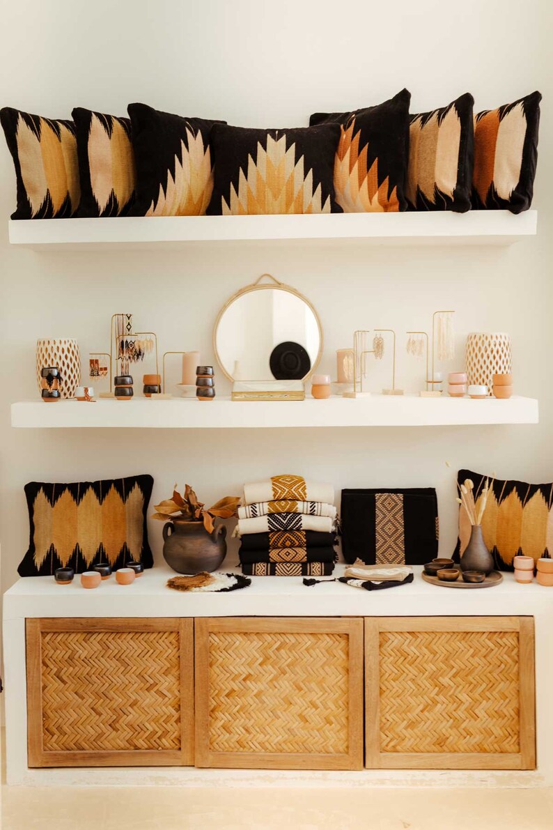 May include: A white shelf unit with two shelves, featuring a variety of home decor items, including woven pillows, a round mirror, candles, and decorative vases. The bottom shelf has a wooden cabinet with three woven doors.