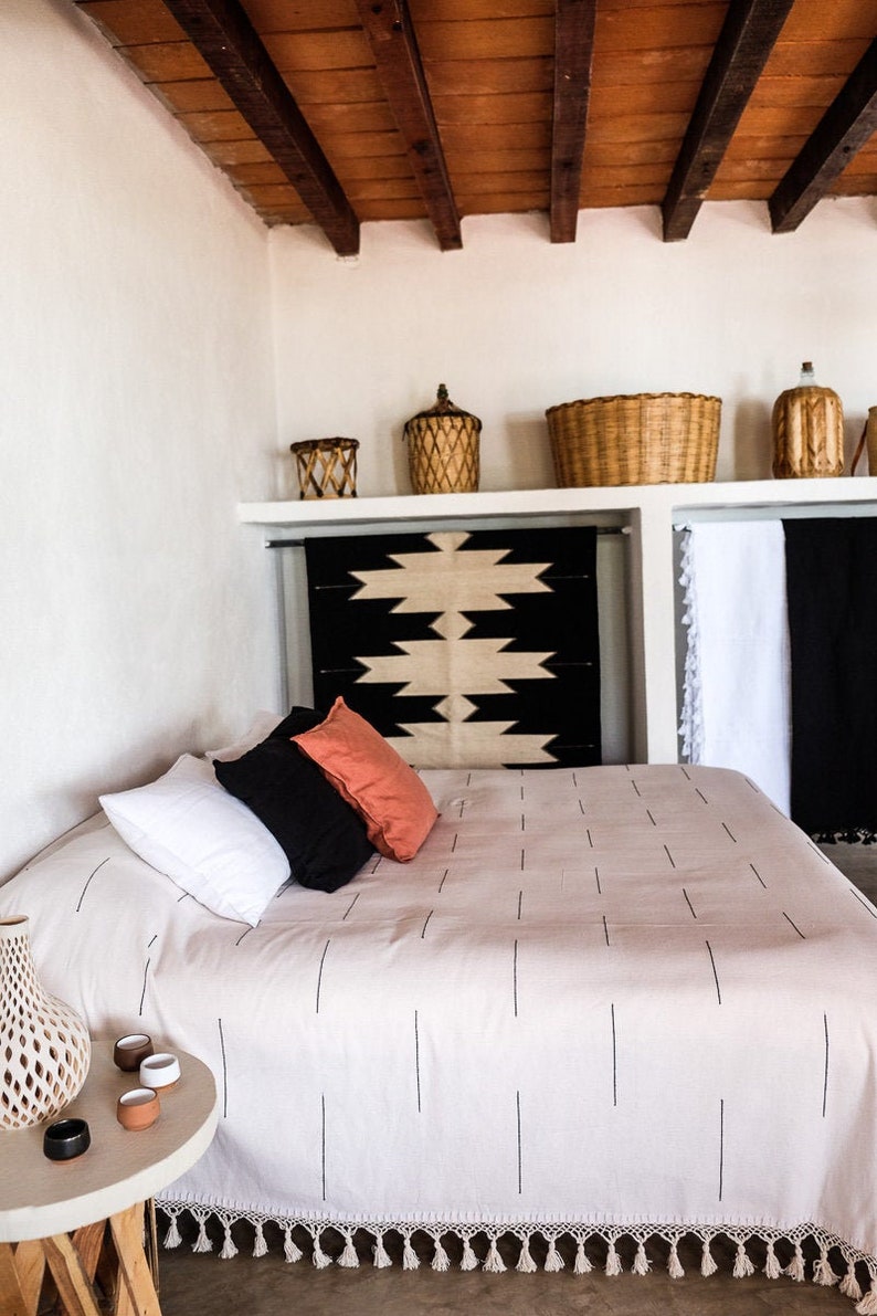 Puede incluir: Una cama blanca con una manta de patchwork blanco y negro y una cabecera con estampado blanco y negro. La cama tiene un ribete de crochet blanco con borlas. Hay tres almohadas en la cama, una blanca, una negra y una naranja.