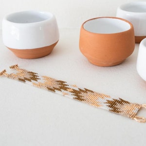 May include: A white and gold beaded bracelet with a geometric pattern. The bracelet is lying on a white surface next to four small ceramic bowls.