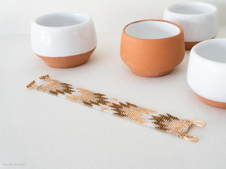 May include: A white and gold beaded bracelet with a geometric pattern. The bracelet is lying on a white surface next to four small ceramic bowls.