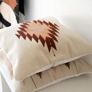 Puede incluir: Dos almohadas de sof&aacute; beige con un patr&oacute;n geom&eacute;trico marr&oacute;n y melocot&oacute;n. Las almohadas est&aacute;n apiladas una encima de la otra.