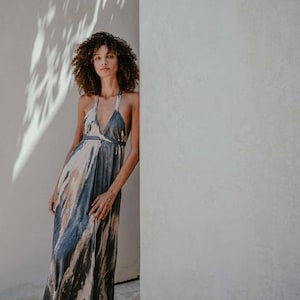 May include: A woman wearing a long, flowing, blue and beige tie-dye dress with a halter neckline. She is standing next to a white wall with a shadow of a tree on it.