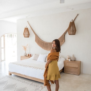 Pode incluir: Uma mulher com um vestido amarelo e um xale castanho est&aacute; de p&eacute; num quarto com uma cama branca, um tapete branco e uma mesa de cabeceira de madeira. O quarto tem um teto branco com um ventilador de teto e duas lumin&aacute;rias penduradas.