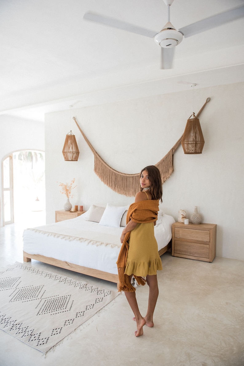 Pode incluir: Uma mulher com um vestido amarelo e um xale castanho est&aacute; de p&eacute; num quarto com uma cama branca, um tapete branco e uma mesa de cabeceira de madeira. O quarto tem um teto branco com um ventilador de teto e duas lumin&aacute;rias penduradas.