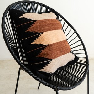 May include: A black woven pillow with a geometric pattern in brown, beige, and black sits on a black metal chair with a circular back.