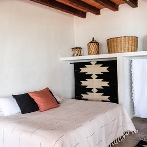Puede incluir: Una cama blanca con un edred&oacute;n blanco con rayas negras y flecos blancos. Al pie de la cama, una manta de patr&oacute;n negro y blanco. Hay dos almohadas en la cama, una negra y otra roja.