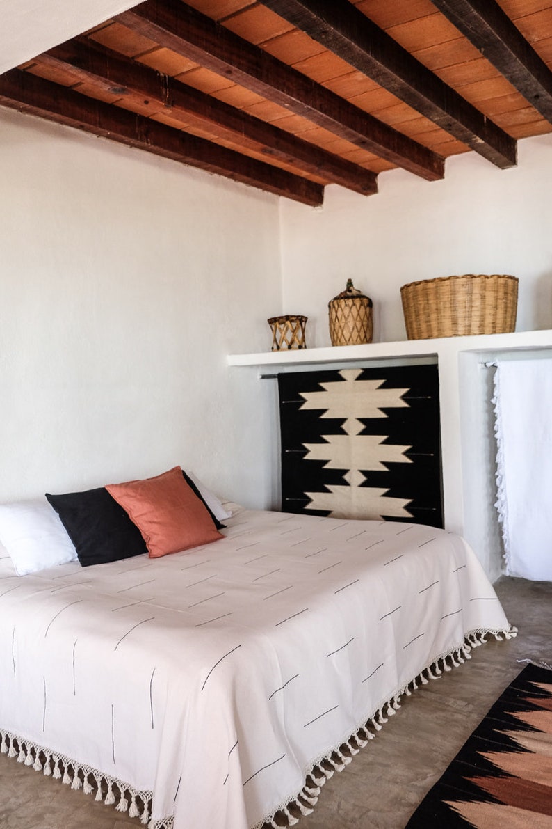Puede incluir: Una cama blanca con un edred&oacute;n blanco con rayas negras y flecos blancos. Al pie de la cama, una manta de patr&oacute;n negro y blanco. Hay dos almohadas en la cama, una negra y otra roja.