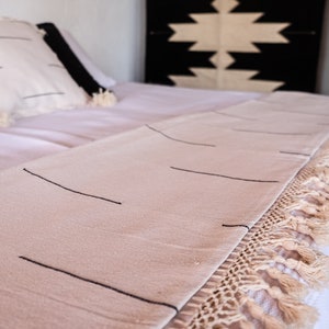 Puede incluir: Una manta de tiro de color crema con rayas negras y un borde de flecos. La manta est&aacute; sobre una cama blanca con una cabecera de patrones blanco y negro.