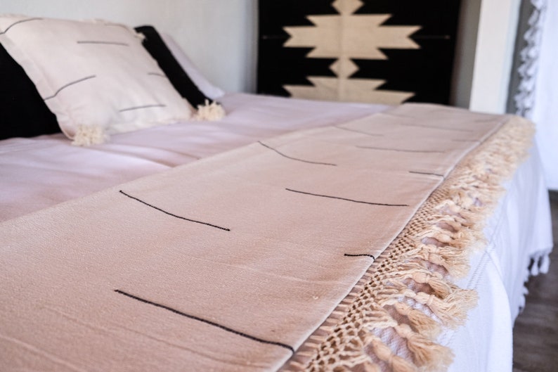 Puede incluir: Una manta de tiro de color crema con rayas negras y un borde de flecos. La manta est&aacute; sobre una cama blanca con una cabecera de patrones blanco y negro.