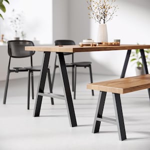 Op de afbeelding: Een houten eettafel met zwarte A-vormige metalen poten en een bijpassende houten bank. De tafel is gedekt met een wit bord, een kom met noten en een vaas met gedroogde bloemen.