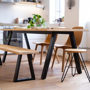 May include: A dining set featuring a long rectangular wooden table with black metal A-frame legs, a matching wooden bench with black metal legs, two tan moulded chairs, and a small round wooden stool with thin black metal hairpin legs.