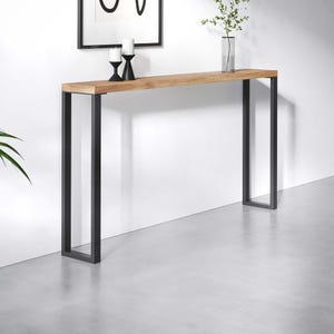 Può includere: Un tavolo consolle alto e stretto con un piano rettangolare in legno color marrone chiaro con venature a vista, sostenuto da due gambe in metallo nero a forma di U. Il tavolo presenta un design lineare pulito con una superficie piana.
