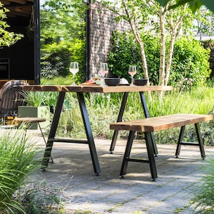 May include: A wooden outdoor dining set with a rectangular table and a bench. The table and bench have a natural wood finish and black metal legs. The set is placed on a stone patio, surrounded by greenery and trees.