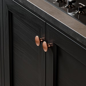 May include: Two copper cabinet knobs are mounted on a black wooden cabinet door. The knobs are round and have a slightly raised centre.