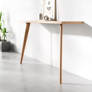 Puede incluir: Una consola de madera clara con una tapa larga y estrecha y dos patas inclinadas. La consola está colocada contra una pared blanca y tiene un diseño simple y moderno.