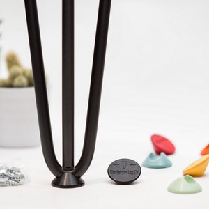 Op de afbeelding: Zwarte metalen haarspeldpoten met een zwarte rubberen voet en een zwarte sticker met de tekst "The hairpin Leg Co.". Er zijn ook verschillende gekleurde rubberen vloerbeschermers.