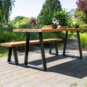 May include: A rectangular outdoor dining table and a matching bench, both constructed from thick, natural-toned wooden planks. The furniture features sturdy, black metal A-frame legs. The table surface holds two wine glasses and two dark bowls.