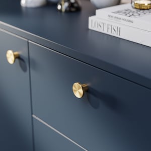 May include: Two gold-toned round cabinet knobs with a brushed finish are mounted on a dark blue cabinet door. The knobs are slightly raised from the surface of the door.