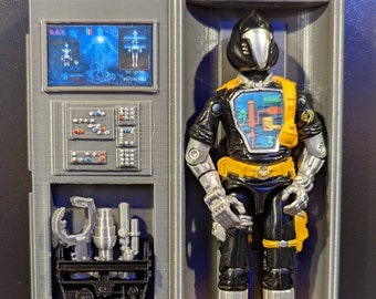 May include: A black and silver action figure with a yellow belt and a black hooded helmet stands in a gray and black display case. The case has a screen with the text "DATA" and "WEAPON SYSTEMS" and a panel with buttons and switches. The figure is holding a silver weapon in its right hand.