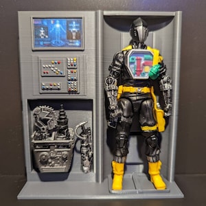 May include: A gray plastic display stand with a black and yellow action figure inside. The figure has a black helmet, a black and yellow suit, and a green and blue chest piece. The stand has a screen with the text "Weapon System" and a control panel with buttons.