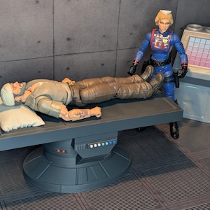 Può includere: Un action figure maschile con barba e benda bianca sulla testa è sdraiato su un lettino medico grigio con un cuscino bianco. Un action figure femminile in uniforme blu si trova accanto a una console informatica grigia con quadrati colorati.