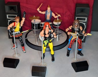 May include: A rock band action figure set featuring five figures on a stage with drums, guitars, and keyboards. The figures are dressed in colorful rock star attire, with microphones and speakers. The backdrop is a red curtain.