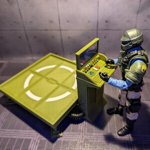 Pu&ograve; includere: Una piattaforma di atterraggio verde e bianca con un pannello di controllo e una action figure verde e blu in piedi accanto ad essa.