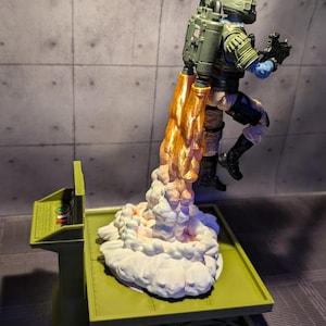Pu&ograve; includere: Una action figure verde e bianca con uno zaino a razzo si lancia da una piattaforma verde. Lo zaino a razzo sta sparando fiamme arancioni e fumo bianco.