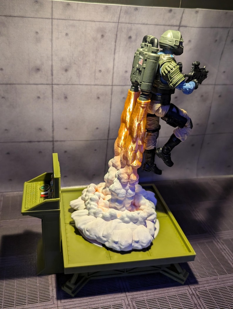 Pu&ograve; includere: Una action figure verde e bianca con uno zaino a razzo si lancia da una piattaforma verde. Lo zaino a razzo sta sparando fiamme arancioni e fumo bianco.