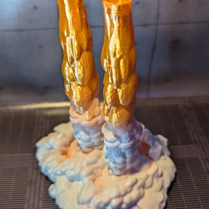 May include: A 3D printed model of a rocket launch with two orange exhaust plumes and a white cloud base.