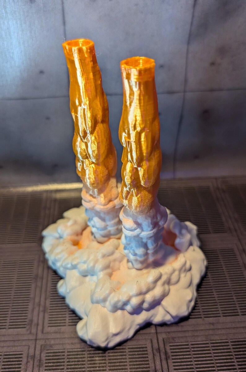 May include: A 3D printed model of a rocket launch with two orange exhaust plumes and a white cloud base.