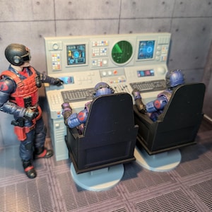 May include: Action figures of soldiers in a command center. One figure in red and black stands near a control panel with screens and buttons. Two figures in blue and black sit in chairs facing the panel. The scene is set in a gray room with a patterned floor.