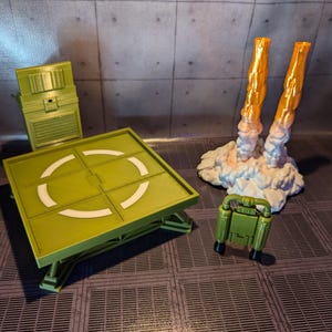 Pu&ograve; includere: Un'area di atterraggio giocattolo verde con un design circolare bianco, un pannello di controllo verde e un jetpack verde con fiamme arancioni.