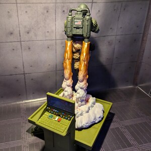 Pu&ograve; includere: Una action figure verde e marrone con uno zaino a razzo e una piccola bandiera americana sul retro. La figura &egrave; in piedi su una nuvola bianca con un pannello di controllo verde e marrone in primo piano.
