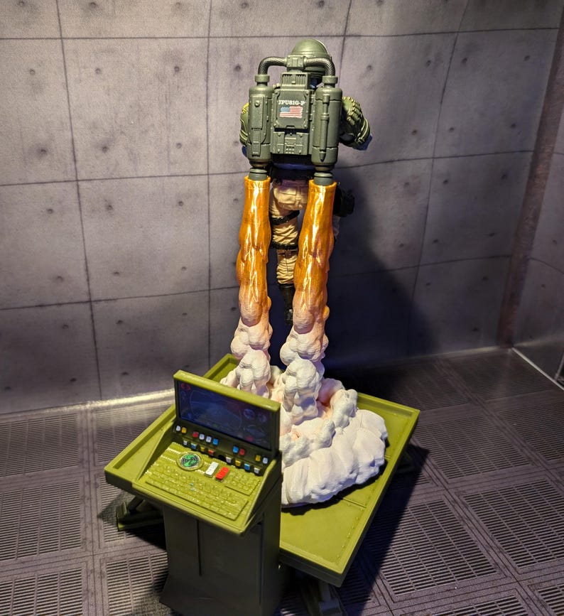 Pu&ograve; includere: Una action figure verde e marrone con uno zaino a razzo e una piccola bandiera americana sul retro. La figura &egrave; in piedi su una nuvola bianca con un pannello di controllo verde e marrone in primo piano.