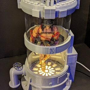 May include: A gray 3D printed model of a science fiction containment unit with a clear glass cylinder. Inside the cylinder is a gray action figure with red straps. The unit has a round base with a gray pipe and a valve.