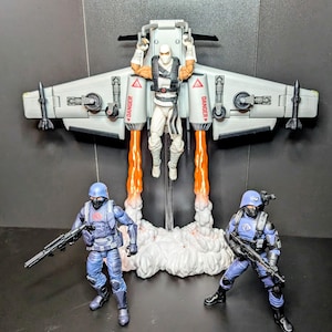 May include: Action figures of a character in a white jumpsuit and a jetpack, with two blue-clad figures holding rifles. The jetpack has orange flame effects and the word "DANGER" on it. The figures are on a black surface.